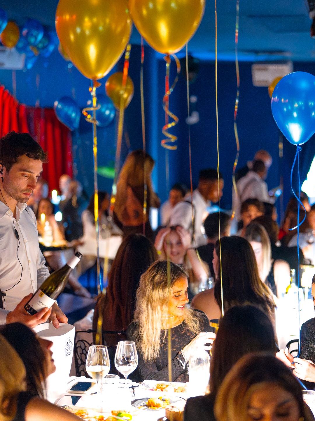 Un cameriere con la bottiglia di vino serve un tavolo sotto i palloncini oro e blu, mentre una ospite sorride