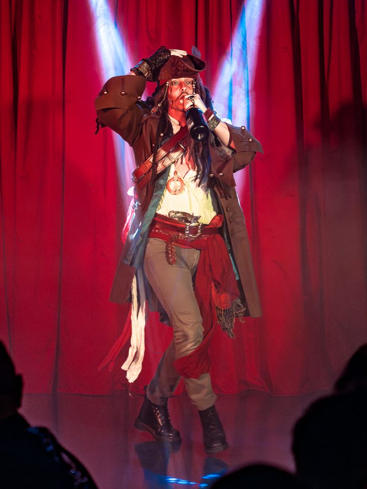 Un performer in costume da pirata sul palco davanti al sipario rosso, illuminato da due fasci di luce